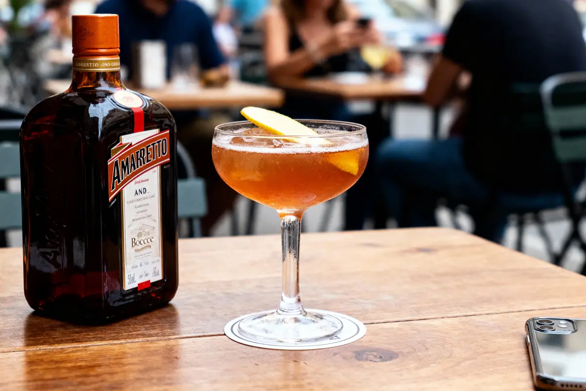 Bouteille d'amaretto et cocktail sur table en bois, terrasse de café floue en arrière-plan.
