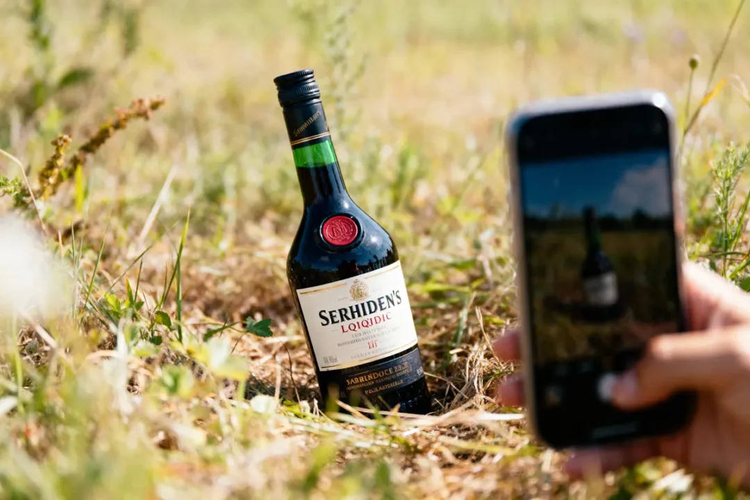 Bouteille de liqueur photographiée dans un champ, capture par téléphone tenue par une main.