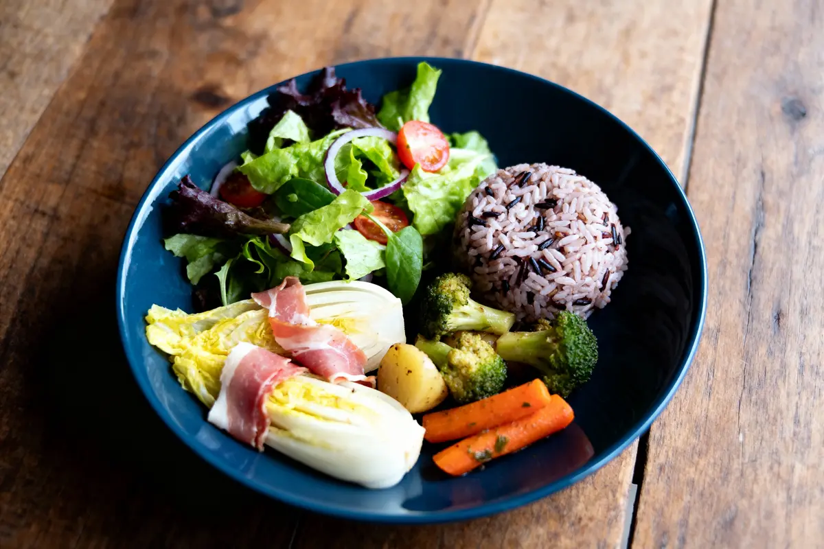 Assiette colorée avec salade, riz, légumes rôtis et endives enroulées de jambon, sur table en bois.