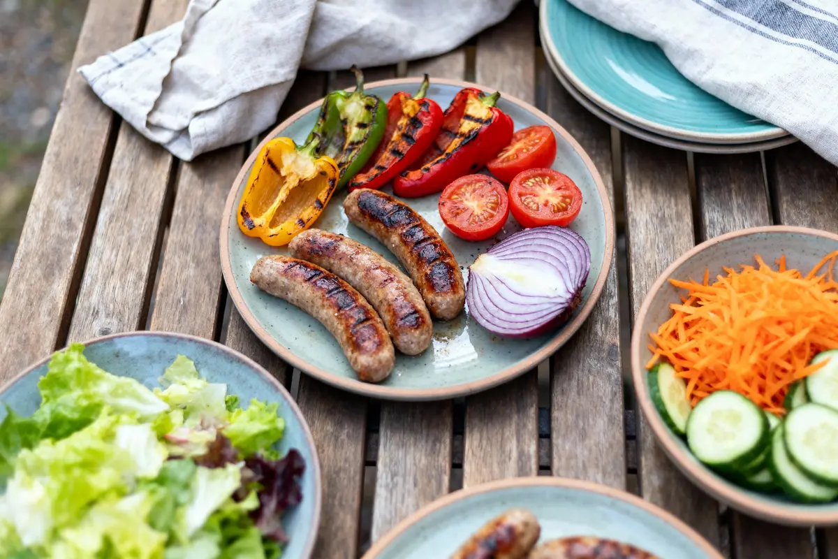 Assiettes de saucisses grillées, légumes, salades variées sur une table en bois extérieure, avec torchon blanc.