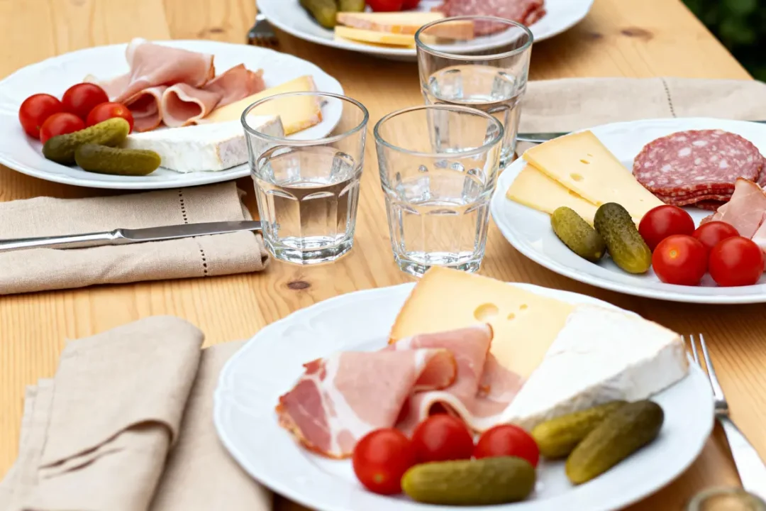 Assiettes de charcuterie, fromage, cornichons, tomates cerises sur une table en bois avec verres d'eau et serviettes.