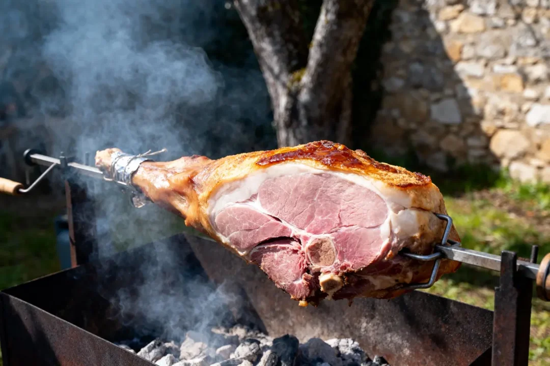 Gigot rôti sur broche au-dessus de charbon, extérieur ensoleillé, fumée visible, arrière-plan de pierre et arbres.