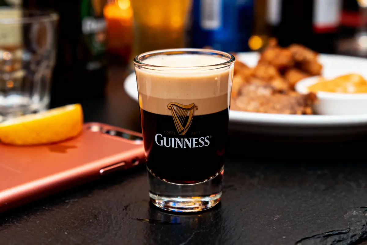 Verre de Guinness sur table, téléphone rose, citron, assiette en arrière-plan. Ambiance de bar.