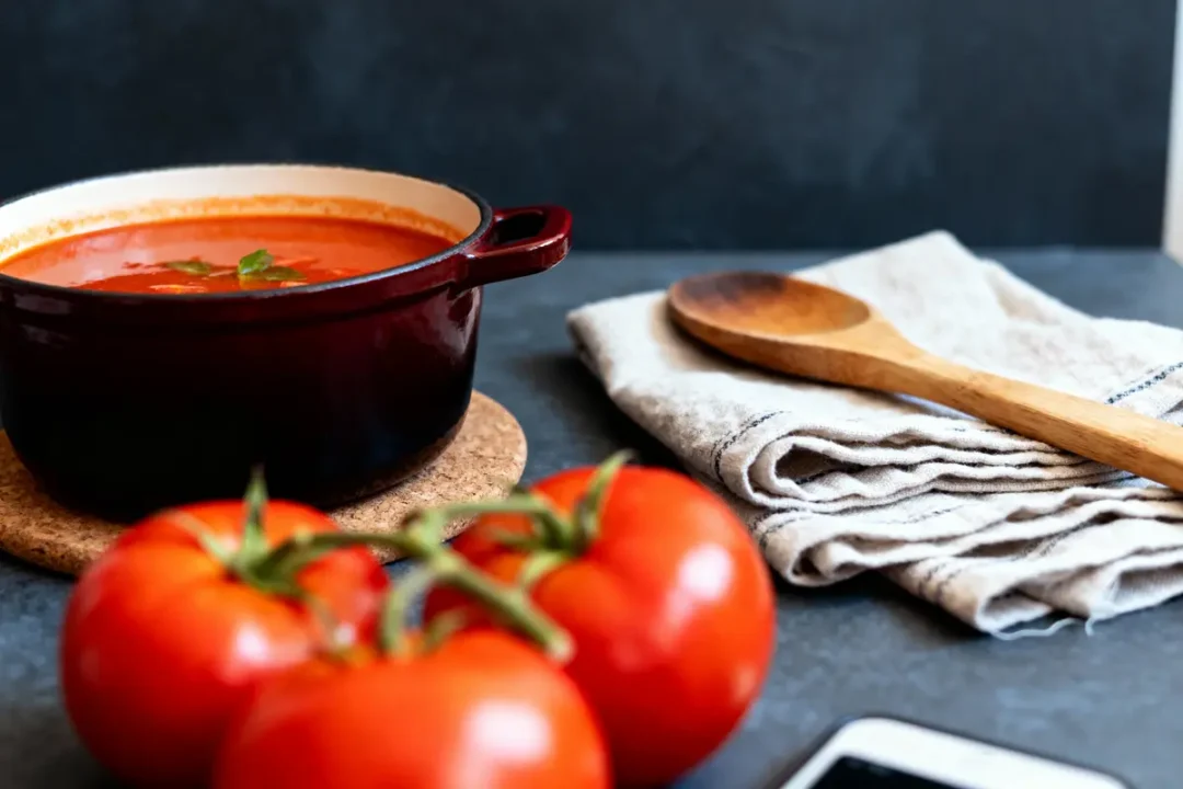 Casserole de soupe tomate sur dessous de plat, torchon plié, cuillère en bois, tomates fraîches au premier plan.