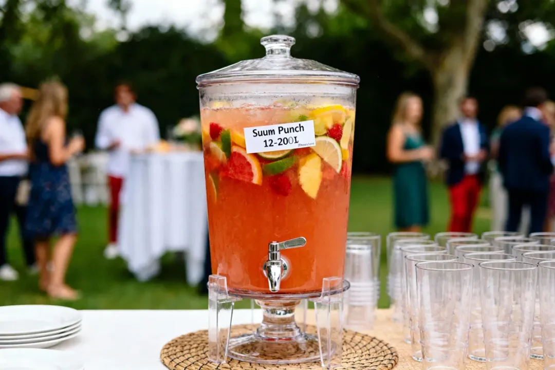 Distributeur de punch fruité avec verres, en extérieur lors d'une réception, invités flous en arrière-plan.